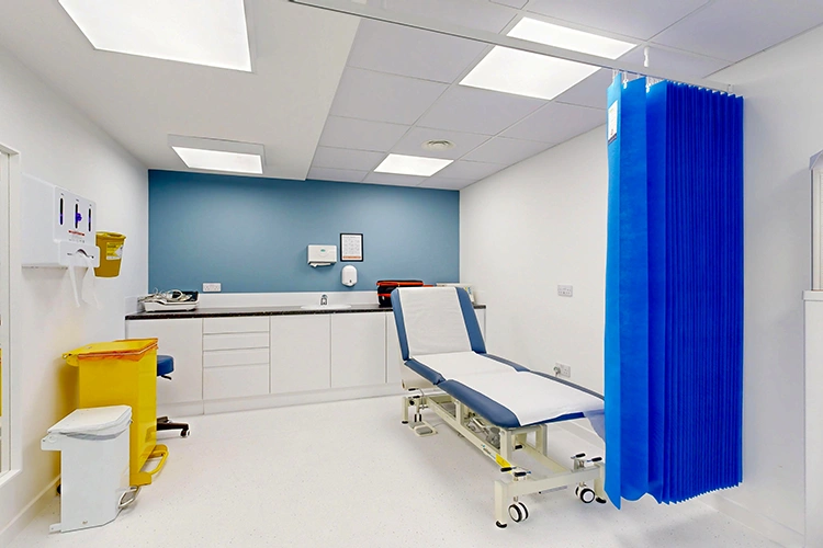 Consultation room at London Private Hospital with adjustable examination couch, blue privacy curtain, digital weighing scale and patient chair.